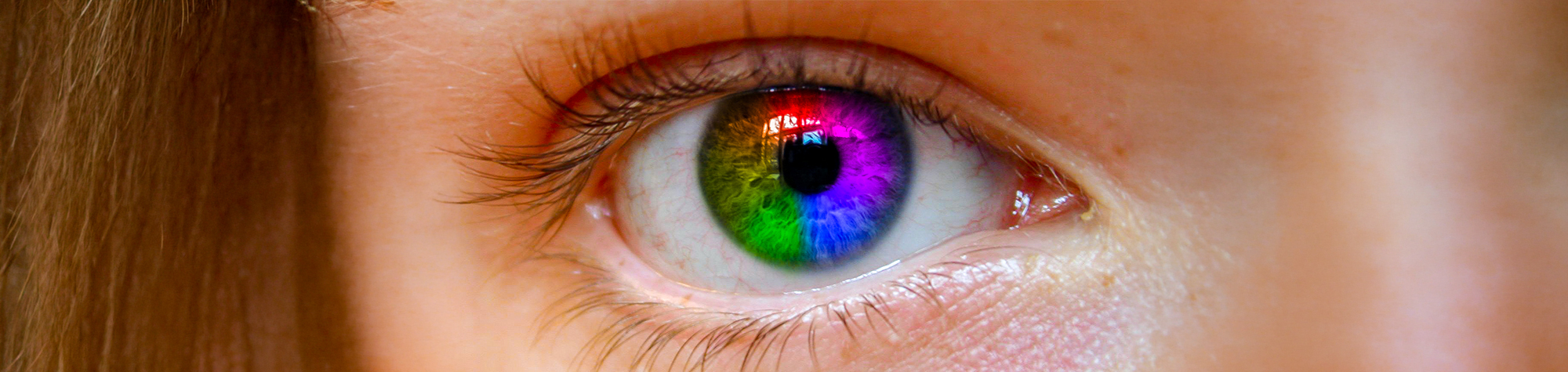 A wide panoramic banner showing a high-detail macro shot of a human eye where the iris has been transformed into a vibrant, seamless rainbow gradient using Photoshop blending modes.
